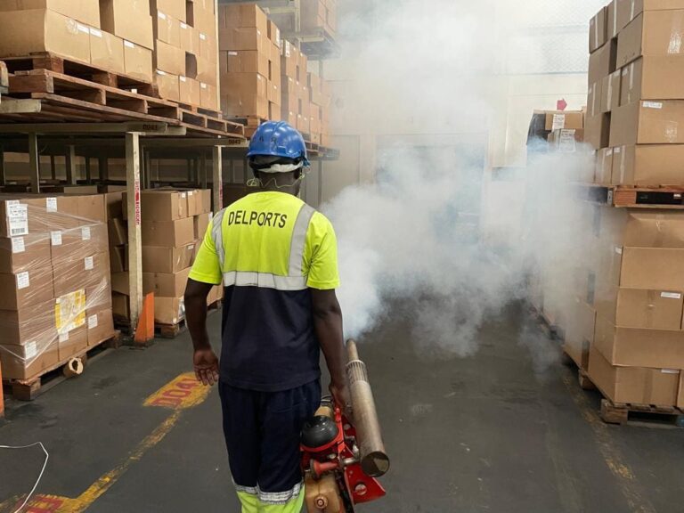 A Delports specialist, equipped with safety gear including a hard hat and protective clothing, uses fogging spray to eliminate tobacco beetles, moths and pathogens in hard-to-reach areas of a tobacco warehouse. This method ensures thorough coverage for hygienic, pest-free environments Delports Pest Control Specialist Safely Applying Fogging Treatment in a Tobacco Warehouse
