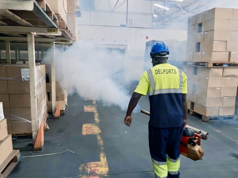 A Delports specialist, equipped with safety gear including a hard hat and protective clothing, uses fogging spray to eliminate tobacco beetles, moths and pathogens in hard-to-reach areas of a tobacco warehouse. This method ensures thorough coverage for hygienic, pest-free environments Delports Pest Control Specialist Safely Applying Fogging Treatment in a Tobacco Warehouse